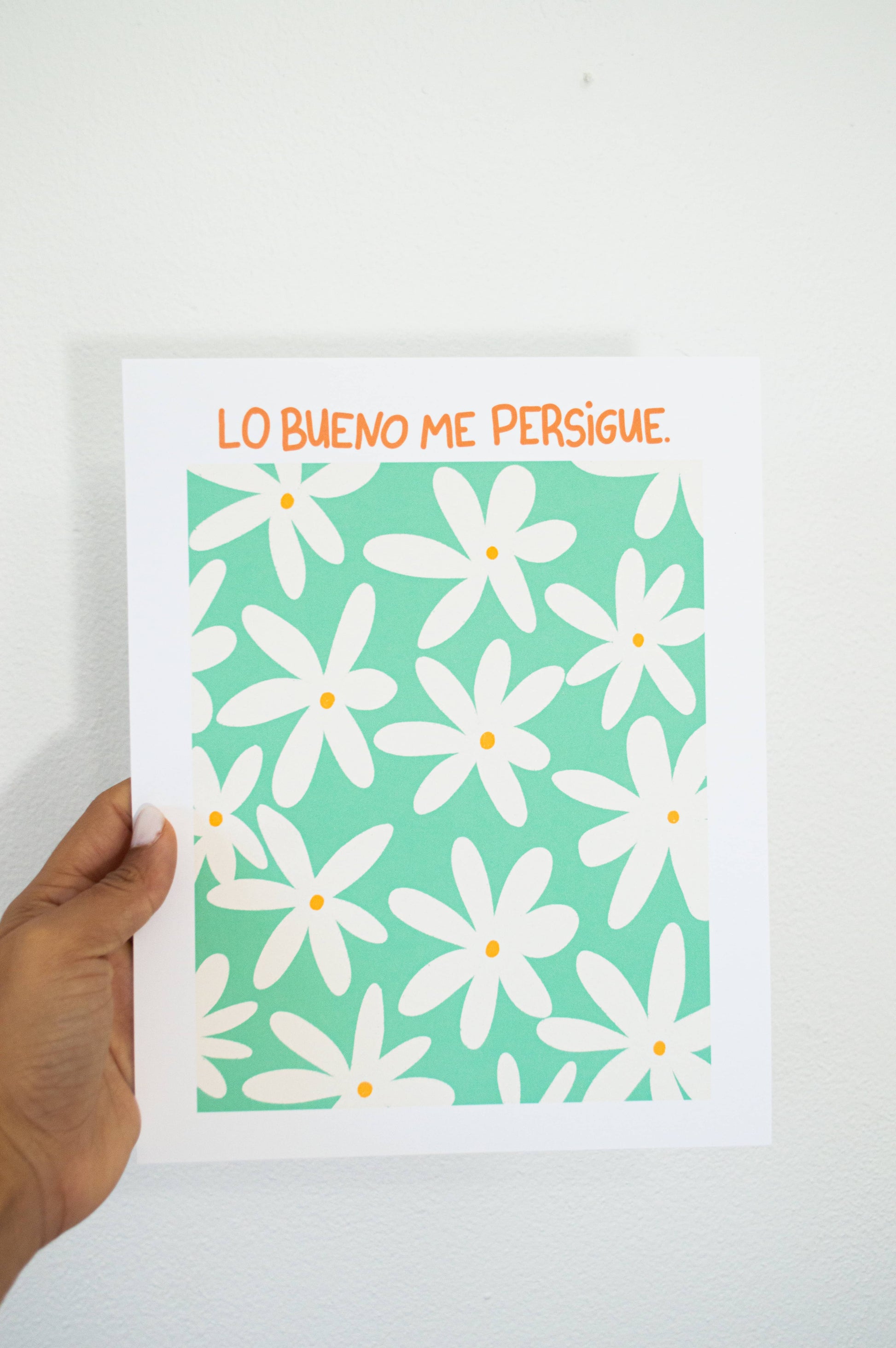 Ilustración con el mensaje "Lo Bueno Me Persigue" en fondo turquesa y flores crema.